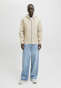 Chaqueta impermeable beige con capucha, cierre completo y dos bolsillos delanteros; combinada con jeans de corte suelto en un tono azul claro y zapatillas grises.