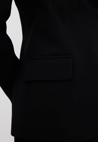Černý blejzr s elegantním designem, hladkým materiálem a obdélníkovou náprsní kapsou po straně, s jemnými švy.