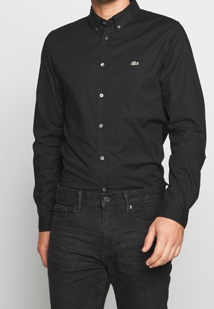 Homme portant une chemise noire ajustée à boutons avec un petit logo crocodile vert sur la poitrine et un jean noir, debout avec une main visible.