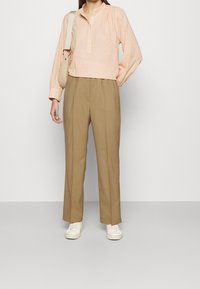 Blouse rose clair à manches longues avec des détails texturés et une patte de boutonnage, associée à un pantalon taille haute beige et des baskets blanches.