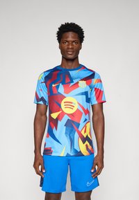 Kék grafikus rövidujjú ing, piros, sárga és sötétkék absztrakt mintákkal, lekerekített nyakkivágással és Barcelona logóval. Kék sorttal viselt.