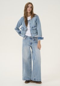 Jachetă din denim albastru deschis cu un design scurt, nasturi în față și două buzunare pe piept. Asortată cu blugi evazați, tot din denim albastru deschis, și pantofi maronii.