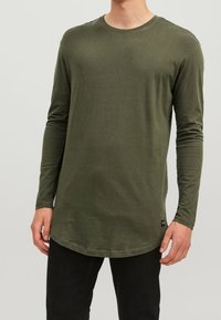 Langärmliges olivgrünes Hemd aus weichem Material, mit einem runden Ausschnitt und einem abgerundeten Saum, kombiniert mit schwarzen Hosen.