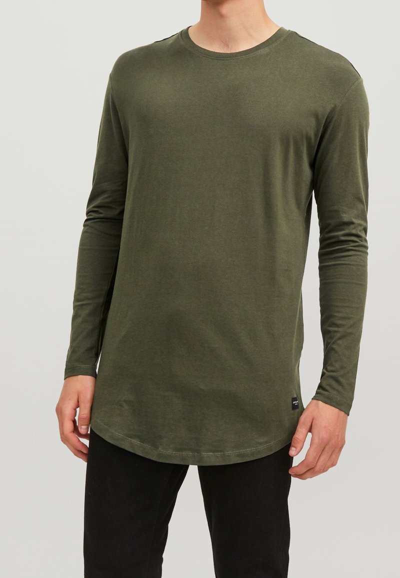 Langärmliges olivgrünes Hemd aus weichem Material, mit einem runden Ausschnitt und einem abgerundeten Saum, kombiniert mit schwarzen Hosen.