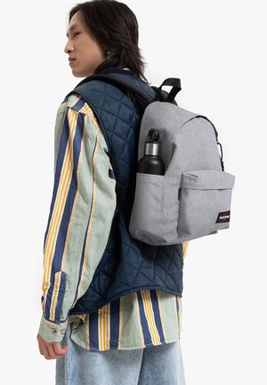 Mochila de tela gris con un bolsillo frontal, llevando una botella de agua negra. El portador viste un chaleco acolchado azul y una camisa de rayas multicolor.