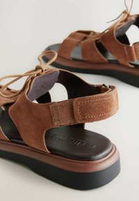 Las sandalias de ante marrón tienen un diseño abierto con correas ajustables y una plantilla acolchada. La suela es oscura y cuenta con una ligera altura de plataforma.