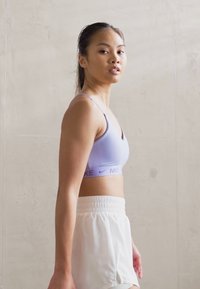 Világos lila sportmelltartó Nike márkajelzéssel, fehér, magas derekú rövidnadrággal párosítva. Simább anyag, testhezálló kialakítás, minimalista stílus.