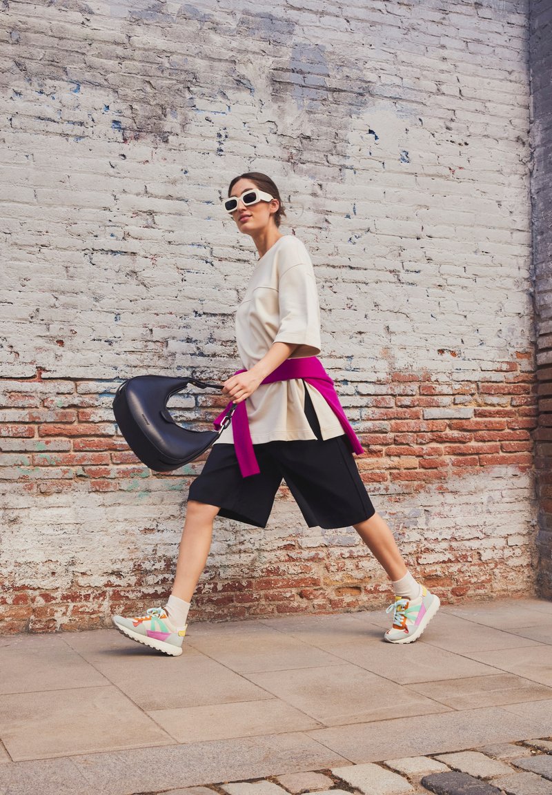T-shirt oversize beige, shorts noirs, robe rose à la taille, et baskets colorées à semelles épaisses, portant un sac à bandoulière noir contre un mur en briques.