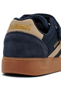 Granatowy zamszowy sneaker z brązowymi akcentami, teksturowaną tkaniną i złotym logo. Posiada gumową podeszwę z wzorzystym bieżnikiem.