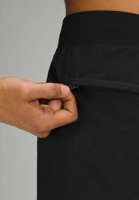 Shorts noirs en tissu lisse et extensible. Une main ouvre une poche latérale discrète à l'aide d'une fermeture éclair noire et d'un design épuré.