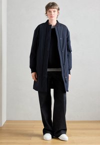 Tmavomodrý dlhý kabát so stojacím golierom, zapínaním na cvoky a bočnými vreckami, oblečený cez čierny sveter a pruhovanú košeľu. Tmavé široké nohavice a biele tenisky.