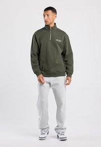 Sudadera verde oliva con cuello con cremallera, con bordado de "Nueva York", combinada con pantalones de chándal grises claros y zapatillas blancas.
