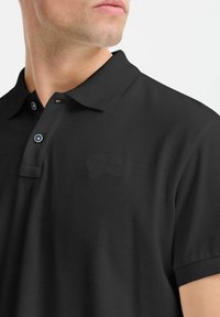 Sort polo skjorte, korte ærmer, flad krave, tekstureret stof, to-knap lukning med kontrastfarvede blå knapper, diskret brandlogo på brystet.