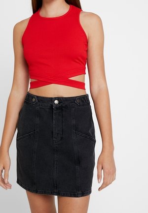 Kvinde iført en ærmeløs rød croptop med krydsede stropper og en sort denimnederdel med høj talje mod en ensfarvet baggrund.
