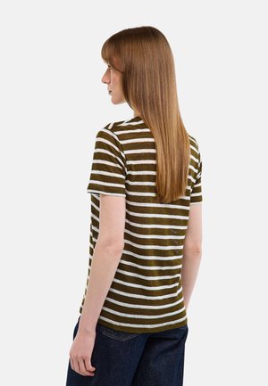 Femme aux cheveux longs, lisses et bruns, portant un t-shirt à manches courtes rayé olive et blanc et un jean bleu foncé, tournée vers la gauche.