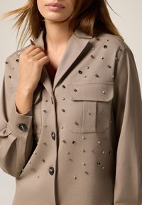 Beige Hemd mit Knopfleiste, Kragen, zwei Taschen und einem Muster aus unterschiedlich großen Strasssteinen auf der Vorderseite und den Ärmeln.