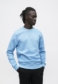 Világoskék pulóver bordázott nyakkivágással és mandzsettával, fehér hímzett "LES DEUX" felirattal. Fekete nadrággal viselve. Sima textúrájú.