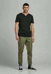 Young man stands facing forward wearing a black short-sleeve shirt, olive cargo pants, and black high-top sneakers with white laces.