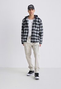 Schwarz-weiß kariertes Flanellhemd über einem weißen Tanktop, kombiniert mit beigen Cargo-Hosen und schwarzen Sneakers. Graue Kappe auf dem Kopf.