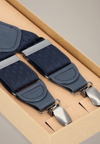 Granatowe szelki z teksturowaną tkaniną, srebrnymi metalowymi klipsami i skórzanymi akcentami, zaprezentowane w kartonowym pudełku.