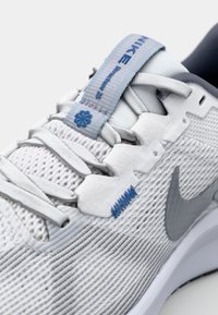 Nike-sneakers med en pustende hvit mesh-overdel, glatte grå detaljer, blå merkevare og et særegent snøresystem.