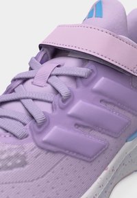 Κοντινό πλάνο ενός αθλητικού παπουτσιού λιλά με υφασμάτινα κορδόνια, λουράκι με βέλκρο και υφή συνθετικού επικαλύμματος στο πλάι.