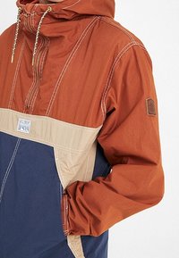 Colorblock anorak met oranje, beige en marineblauwe panelen. Gemaakt van lichtgewicht materiaal met zichtbare stiksels en een logo op de borst.