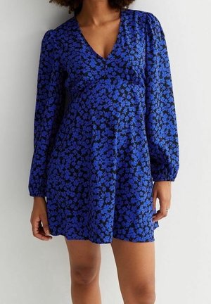 Vrouw die een blauw bloemenjurk draagt met lange mouwen, een V-hals en een losse pasvorm, staand tegen een effen lichte achtergrond.