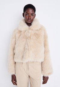 Veste courte en fausse fourrure beige avec un large col et des manches longues, présentant une texture moelleuse et une fermeture à bouton sur le devant.