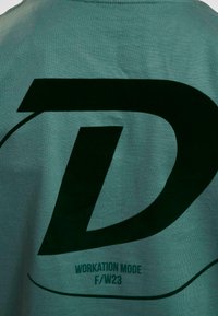 Grønn bomulls t-skjorte med et stort mørkegrønt "D"-logo og teksten "WORKATION MODE F/W23" med mindre skrift nedenfor.