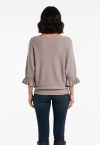 Jasnoszary sweter z fakturowanej tkaniny, o rękawach do połowy przedramienia z falbanami na końcach, noszony z ciemnymi obcisłymi dżinsami.