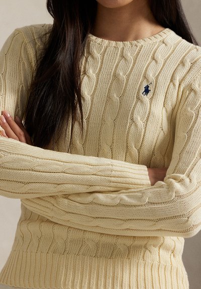Persona con i capelli lunghi e scuri che indossa un maglione beige chiaro a trecce, con le braccia incrociate sul petto.