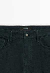 Jean en denim bleu sarcelle foncé avec un design classique à cinq poches, équipé d'une fermeture à bouton métallique et d'une étiquette de marque noire à la ceinture.