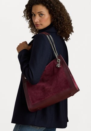 Bordeaux handtas van zacht suède en textuurleer, met een zilveren kettingstrap en een accent van een ontwerperslogo. Ruime, hoekige silhouet.