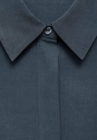 Czarny szary shirt z klasycznym kołnierzykiem, pojedynczym zapięciem na guzik i gładką teksturą materiału. Szwy biegną wzdłuż przodu.