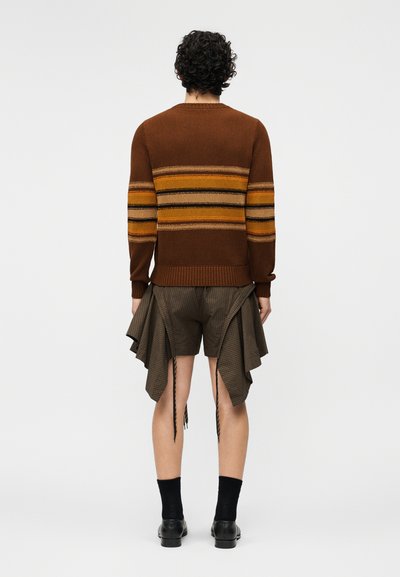 Personne aux cheveux bouclés vue de dos, portant un pull à rayures marron et un short marron à carreaux avec des panneaux de tissu drapés.