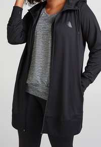 Veste de sport noire avec capuche, dotée d'une fermeture éclair à l'avant, de poches latérales et d'un ourlet côtelé. Portée par-dessus un t-shirt gris et un legging noir.