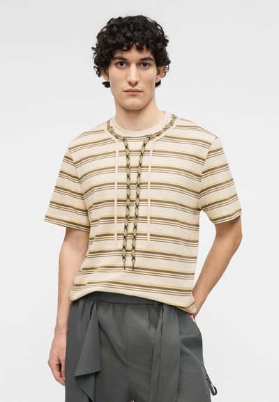 Jeune homme aux cheveux bouclés portant une chemise à manches courtes beige et marron rayée et un pantalon gris ample sur fond blanc.