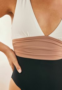Jednodílné plavky s bílým vrškem, béžovým zřaseným středem a černým spodkem. Tkanina má hladkou, elastickou texturu.