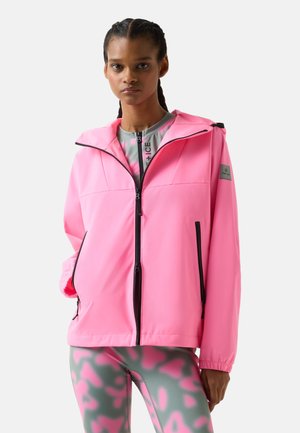 Femme aux cheveux tressés portant une veste zippée rose vif et des leggings gris avec des motifs abstraits roses, debout devant un fond uni.