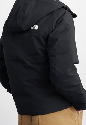 Veste noire à capuche avec le logo "The North Face" sur le haut du dos portée par une personne avec un pantalon beige, mains dans les poches, de dos.