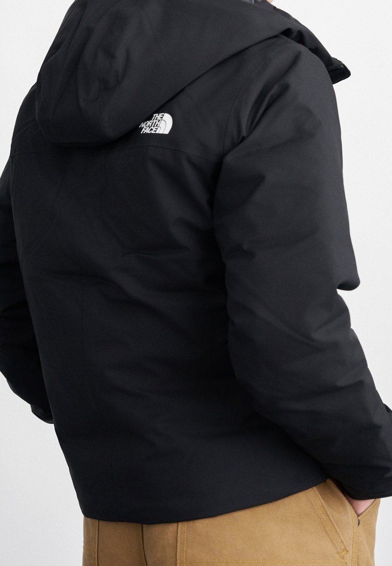 Veste noire à capuche avec le logo "The North Face" sur le haut du dos portée par une personne avec un pantalon beige, mains dans les poches, de dos.