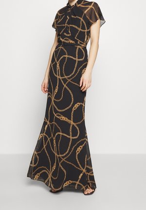 Femme portant une robe longue noire avec un motif chaîne dorée, manches courtes et un col à nouer, debout sur un fond blanc.