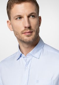 Jasnoniebieska koszula z długim rękawem, wykonana z gładkiego materiału. Posiada kołnierzyk, kieszeń z przodu oraz subtelny wzór. Zbliżenie na górną część ciała.