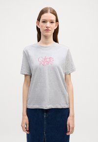 Harmaa puuvillainen t-paita, jossa on pyöreä kaula-aukko ja lyhyet hihat. Edessä vaaleanpunainen "Calvin Klein" -tekstilogo. Liukas rakenne, rento istuvuus.