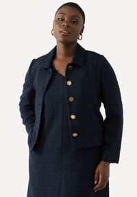 Giacca strutturata blu navy con colletto, caratterizzata da cinque bottoni dorati sulla parte anteriore e maniche lunghe. Indossata sopra un vestito coordinato.