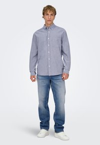 Lyseblå skjorte med knapper og brystlomme, kombineret med falmede blå jeans og hvide sneakers, hvilket skaber et afslappet, lagdelt udseende.