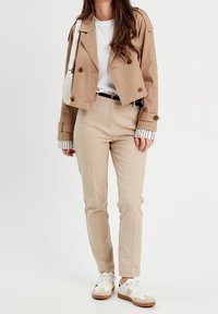 Veste courte beige avec boutons marron, t-shirt blanc, pantalon beige et baskets blanches avec accents marron. Détails de poignets rayés.