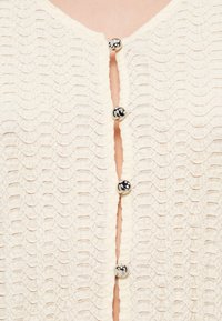 Cardigan de couleur crème avec une texture en motif de vagues, doté de gros boutons ronds noirs et blancs sur le devant et d'un bord festonné.