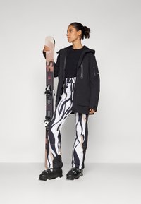 Ski outfit featuring a black jacket, black top, patterned white and black pants, and ski boots. Holding brown snow skis with bindings.